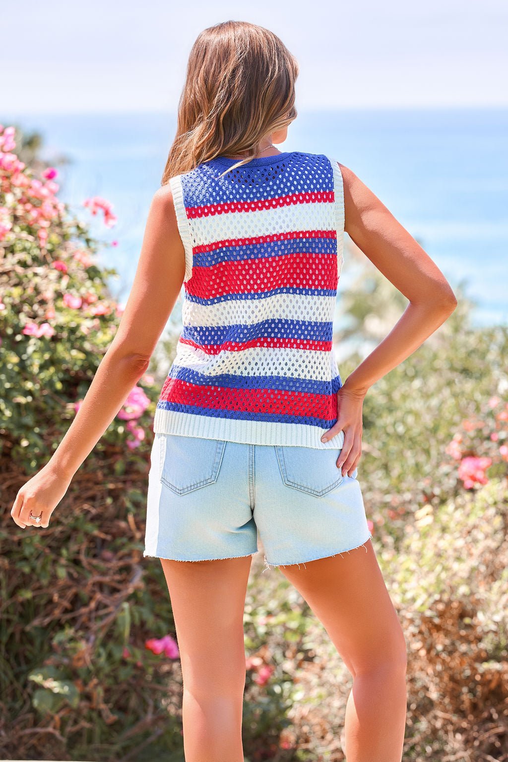 Woman wearing the Stars & Stripes Tie Front Sweater Vest stands outdoors with greenery and an ocean view, facing away in light denim shorts.