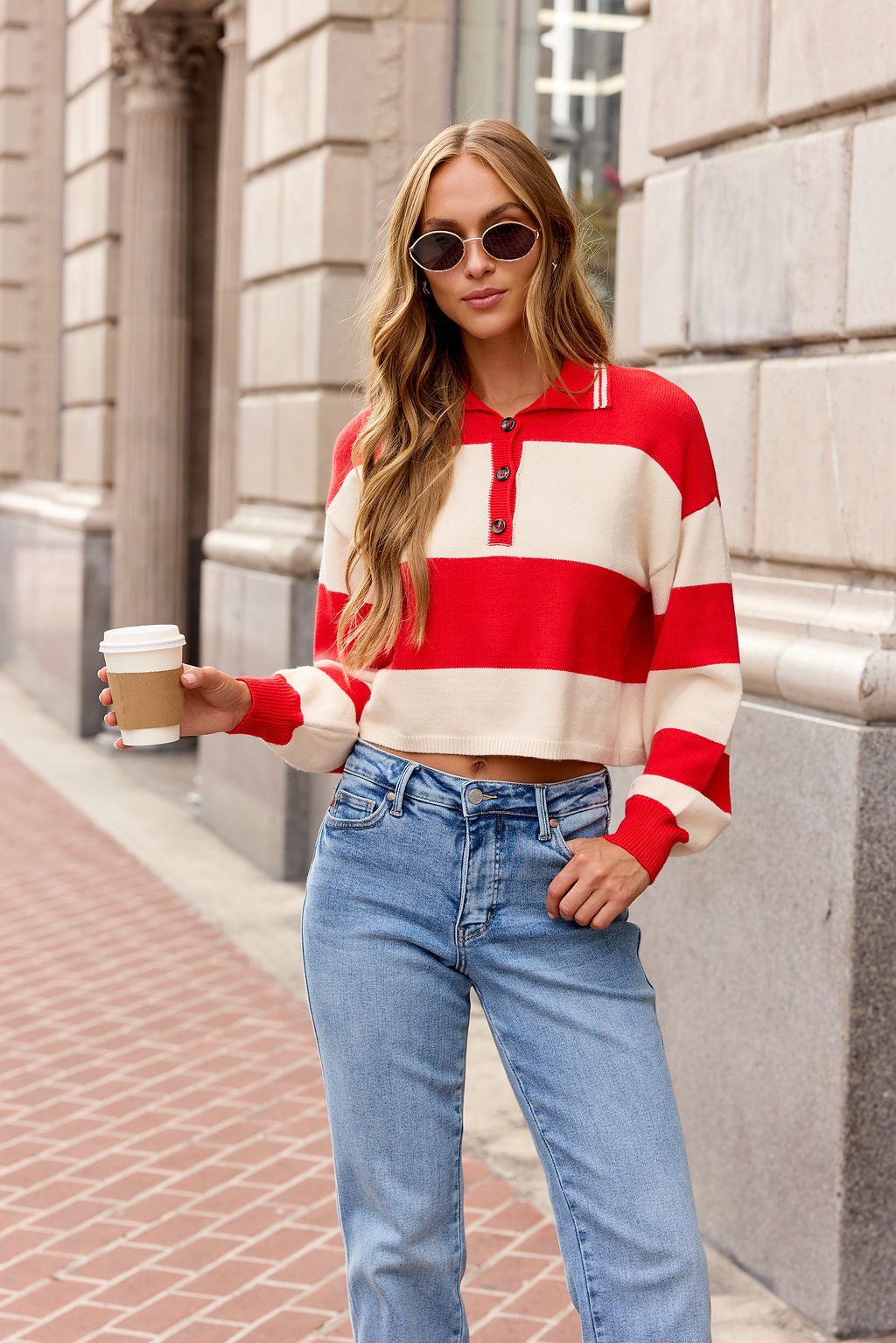 Woman in a red Striped Button Up Sweater with sunglasses and jeans holds coffee on a city sidewalk.