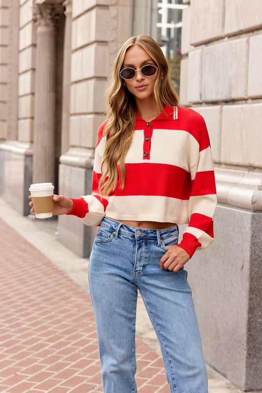 Woman in a red Striped Button Up Sweater with sunglasses and jeans holds coffee on a city sidewalk.
