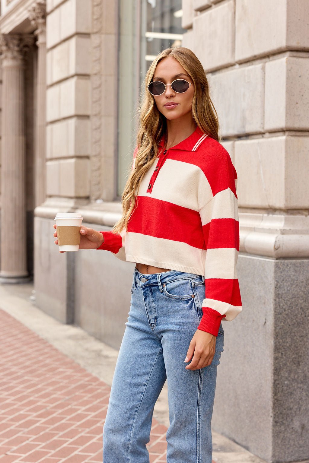 Woman in a Striped Button Up Sweater - Red with jeans holds coffee, wearing sunglasses on a city sidewalk.