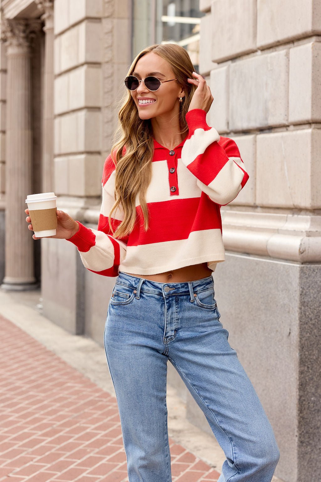Woman in a red Striped Button Up Sweater, jeans, and sunglasses smiles with coffee while walking on the city sidewalk.
