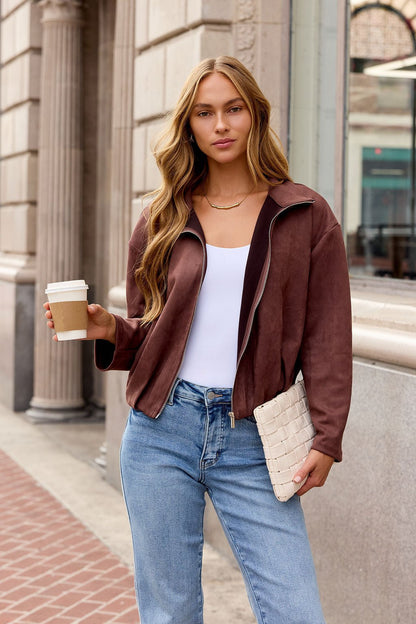 Woman in a brown Suede Zip Up Jacket holds coffee and white clutch on a city sidewalk, looking at the camera.