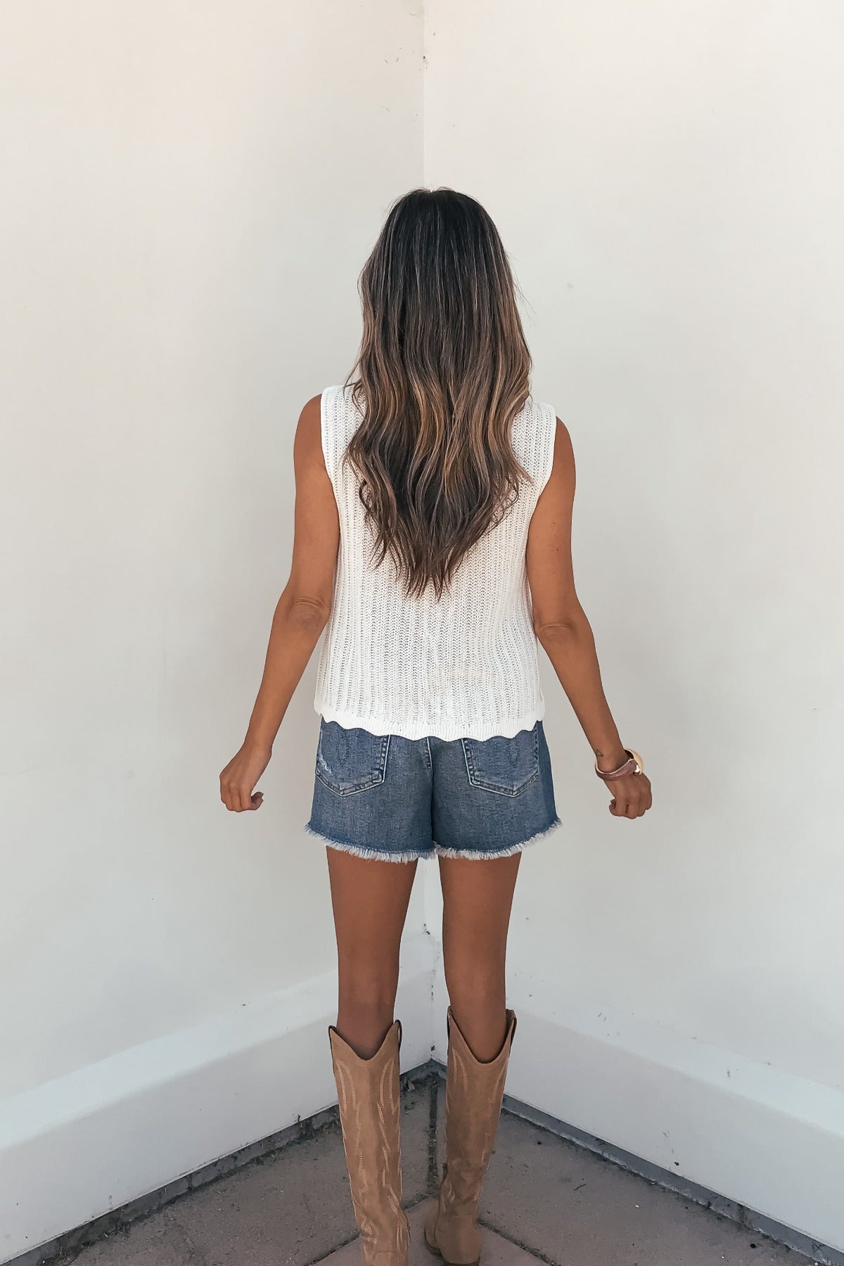 Woman with long wavy hair wears a Sunlace Tie Front Sweater Vest in cream, denim shorts, and tan boots, facing a white wall.