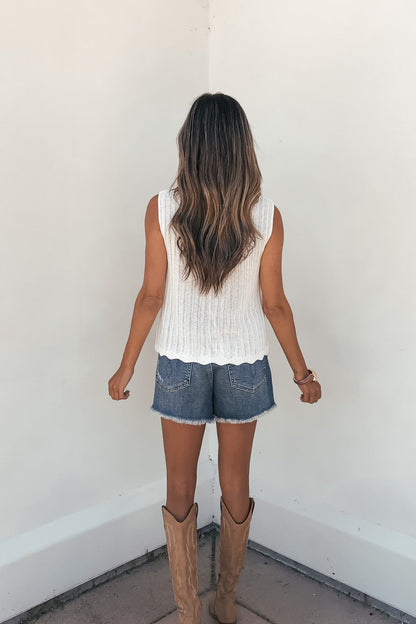 Woman with long wavy hair wears a Sunlace Tie Front Sweater Vest in cream, denim shorts, and tan boots, facing a white wall.