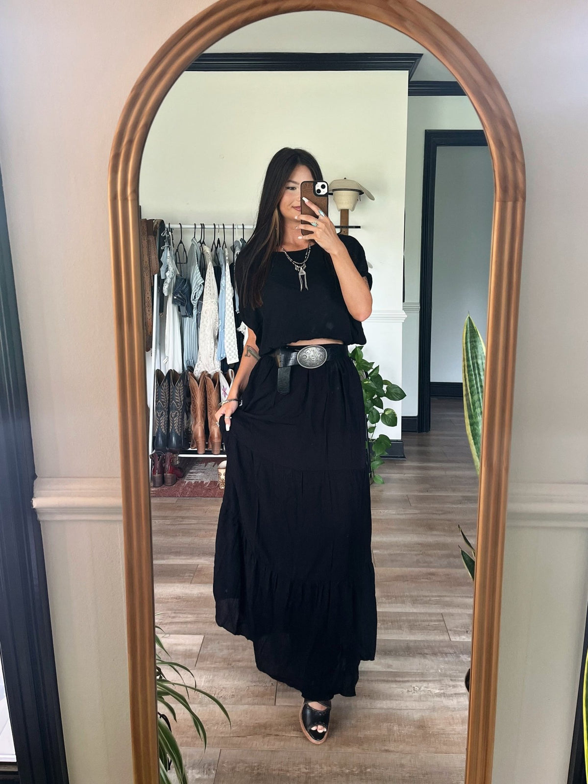 Woman wears the Sunrise Short Sleeve Top & Maxi Skirt Black Set, snapping a mirror selfie in a chic room with plants and hanging clothes.