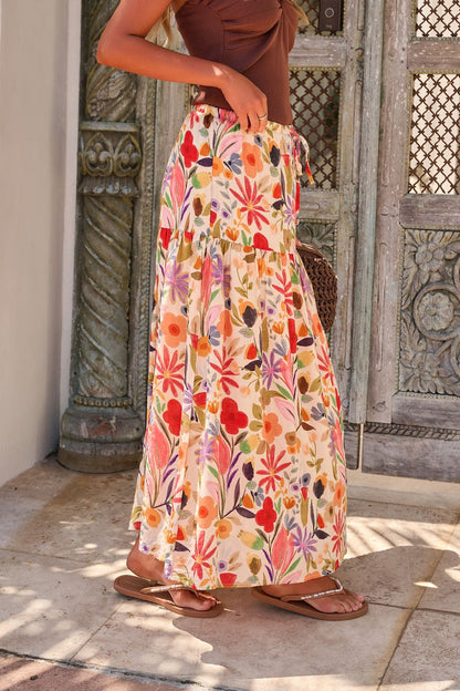 A woman in the Sunset Bloom Multi Print Tiered Maxi Skirt, brown top, and sandals stands by an ornate wooden door.