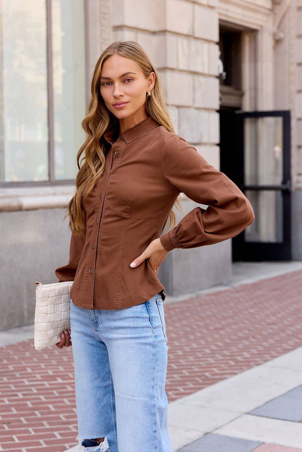 Woman with long wavy hair wears the Sutton Twill Button Down Top in Mocha, light jeans, and holds a white clutch on a city sidewalk.