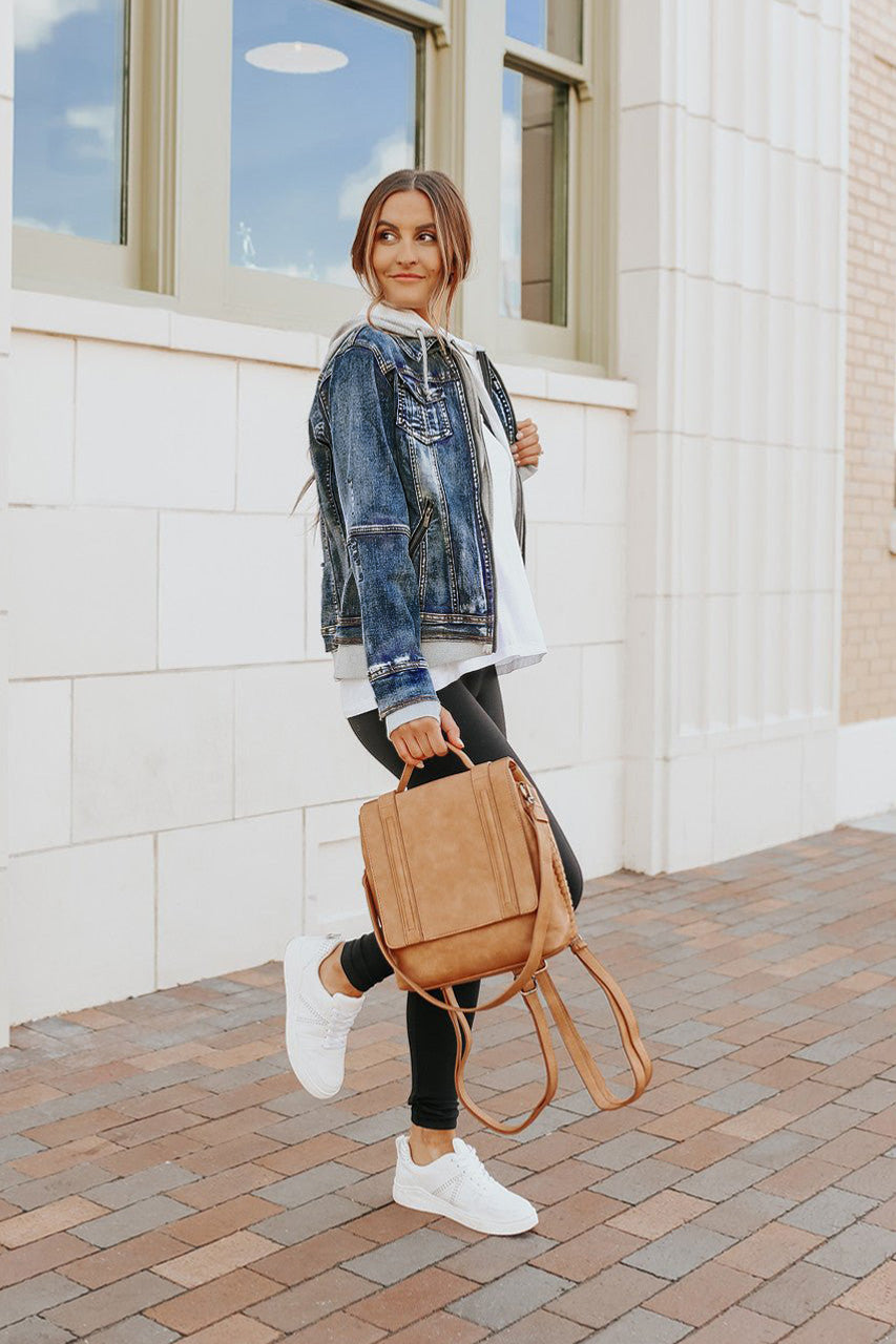 Woman outdoors in a medium wash Denim Jacket with Hoodie, white sneakers, black leggings, and tan backpack.