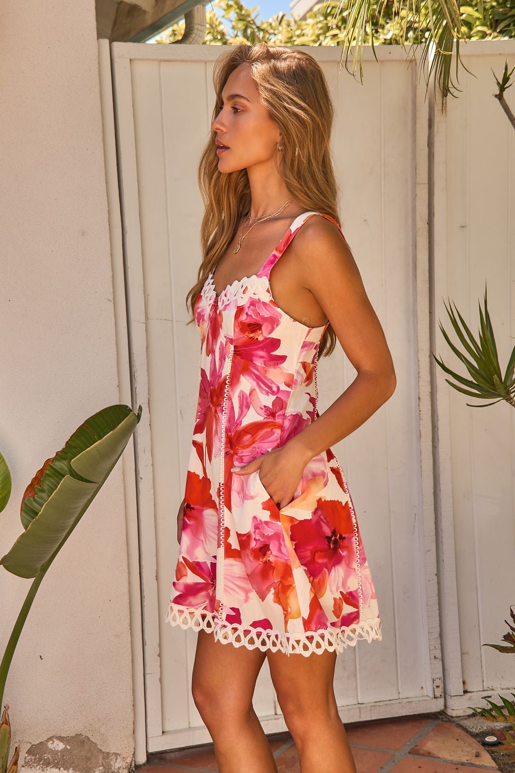 Woman in the Sweet Orchard Print Mini Dress - Pink stands by a white gate, surrounded by greenery, looking off to the side.