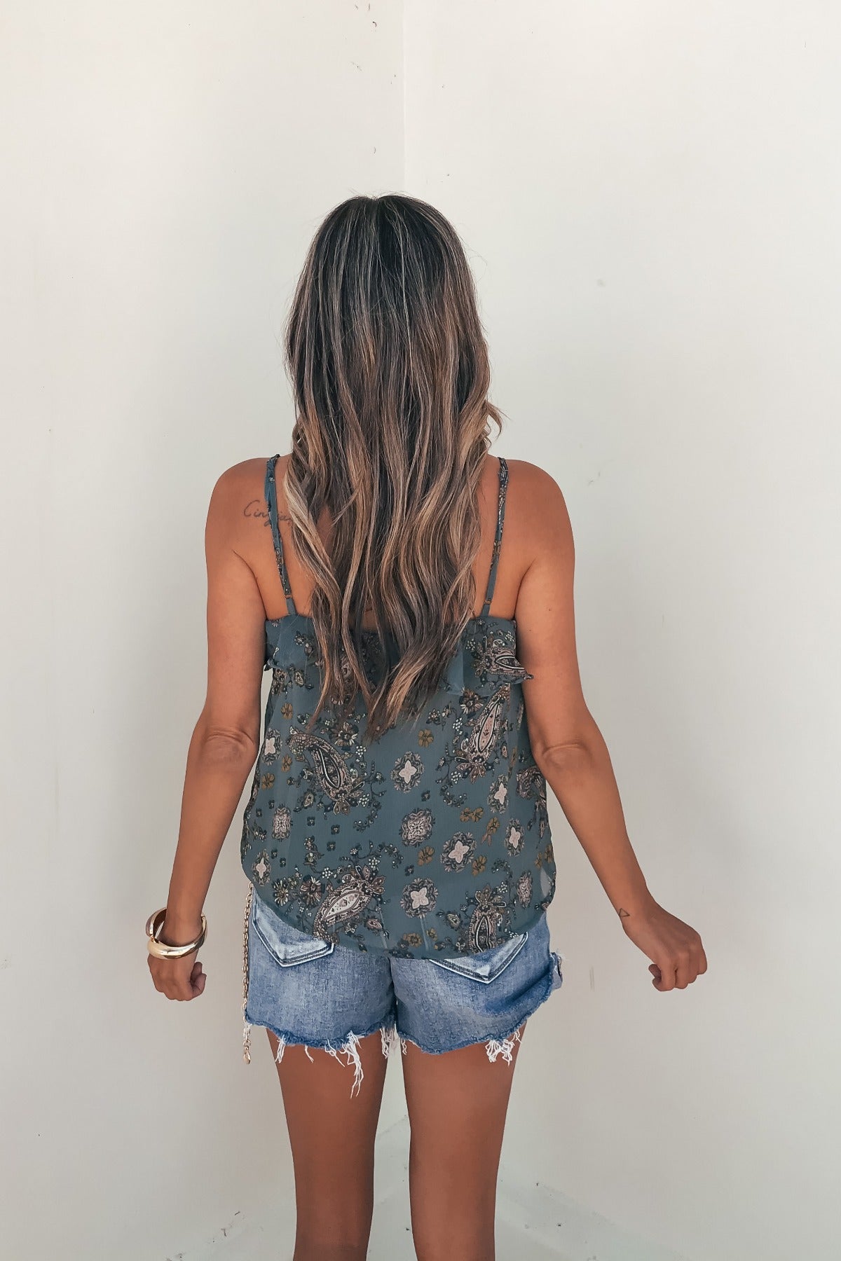 Woman with long wavy hair wears the Teal Paisley Print Ruffled Cami and denim shorts, standing facing a plain white wall.