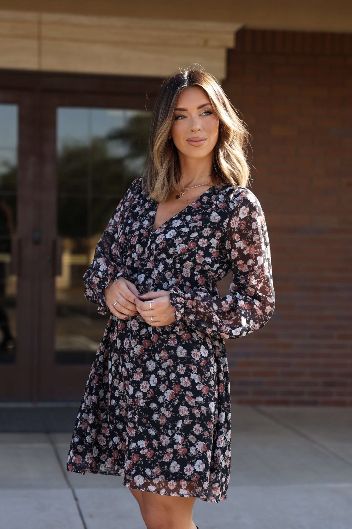 Woman with shoulder-length hair in the Thalia Black Floral Print Mini Dress - FINAL SALE, standing outside by glass doors.