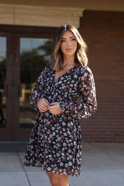 Woman with shoulder-length hair in the Thalia Black Floral Print Mini Dress - FINAL SALE, standing outside by glass doors.