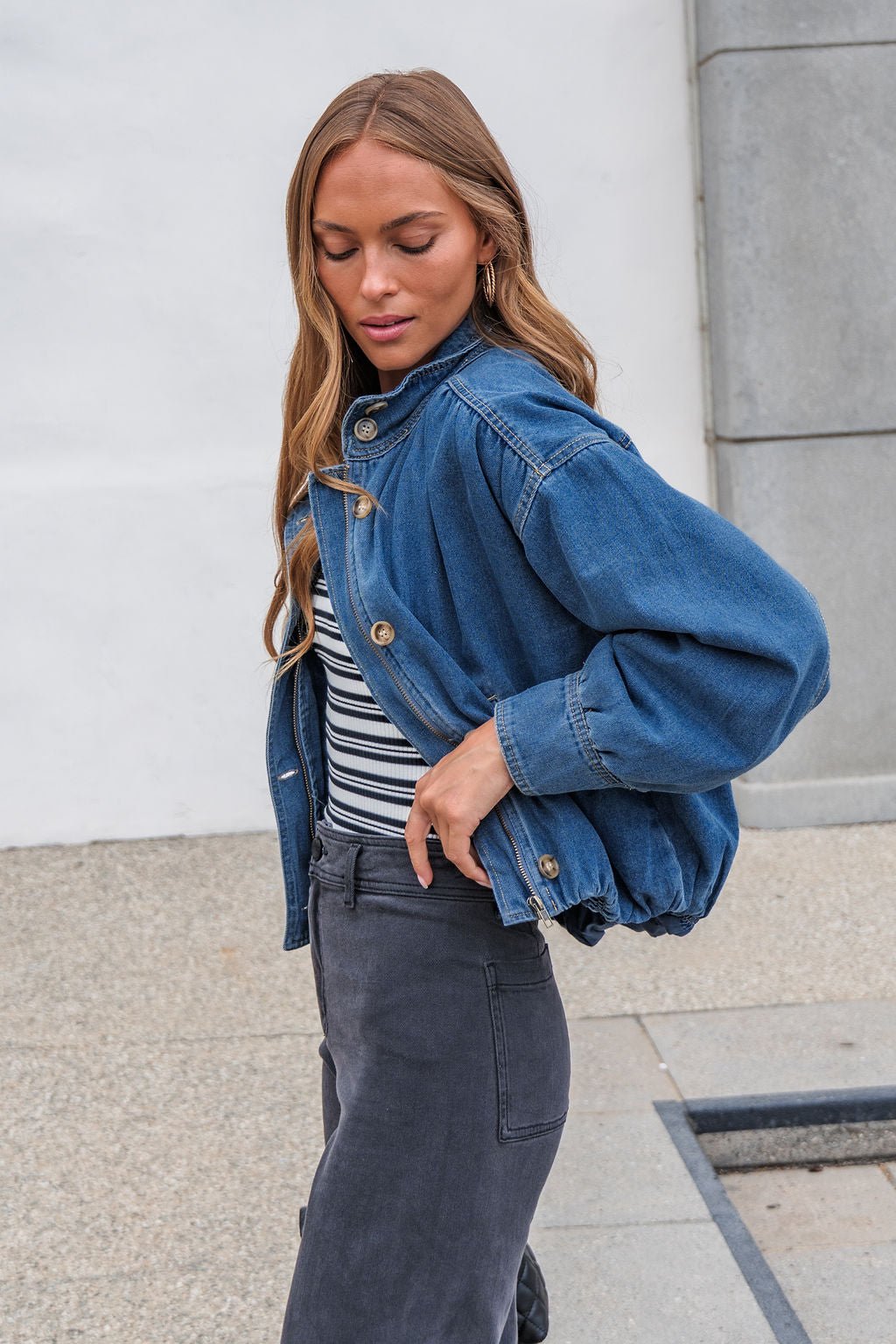 Woman in The Dakota Denim Jacket, striped shirt, and gray pants stands outdoors against a light background.