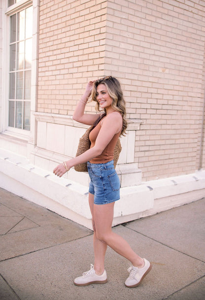 A woman in a brown tank, Thread and Supply Medium Wash Rowan Mid Rise Shorts, and sneakers smiles near a light brick building.