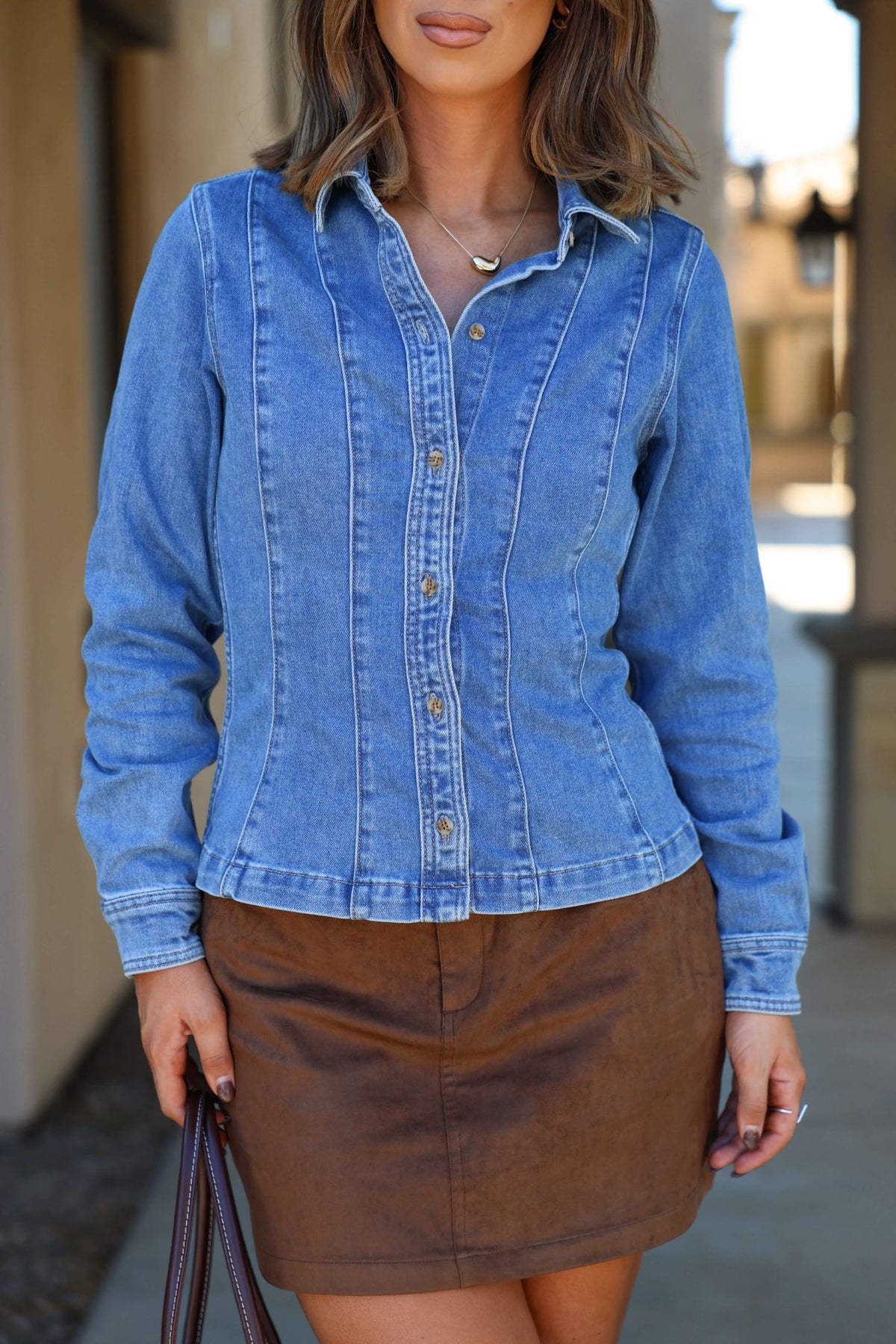 A woman wears the Thread & Supply Button Down Denim Shirt outdoors, pairing it with a brown skirt and holding a bag.