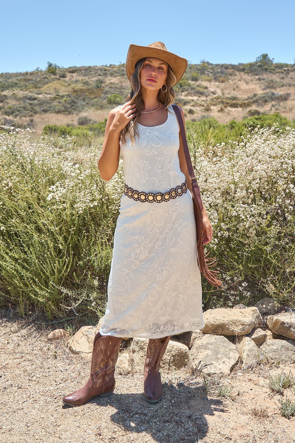 Woman in the Tucson Floral Lace Midi Dress - Ivory stands outdoors with a hat, cowboy boots, floral belt, and brown bag.