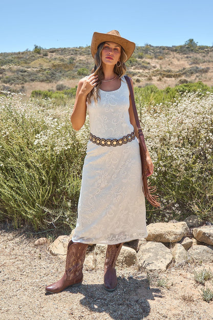 Woman in the Tucson Floral Lace Midi Dress - Ivory stands outdoors with a hat, cowboy boots, floral belt, and brown bag.
