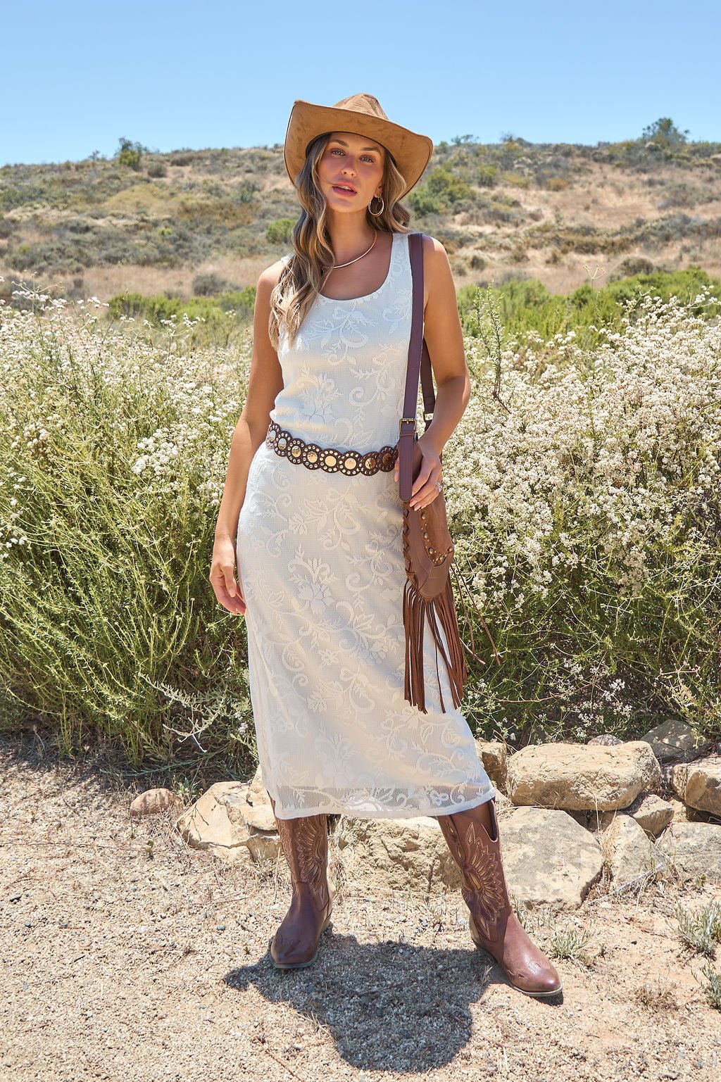 Woman in the Tucson Floral Lace Midi Dress - Ivory stands outdoors, styled with a cowboy hat, boots, and a floral belt amid wildflowers.