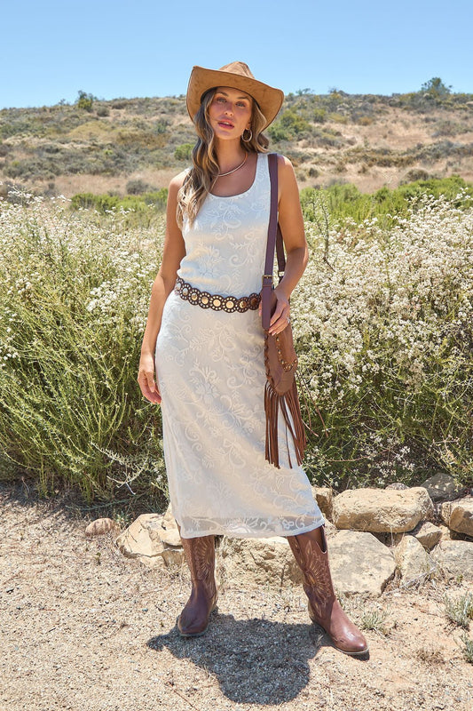 Woman in the Tucson Floral Lace Midi Dress - Ivory stands outdoors, styled with a cowboy hat, boots, and a floral belt amid wildflowers.