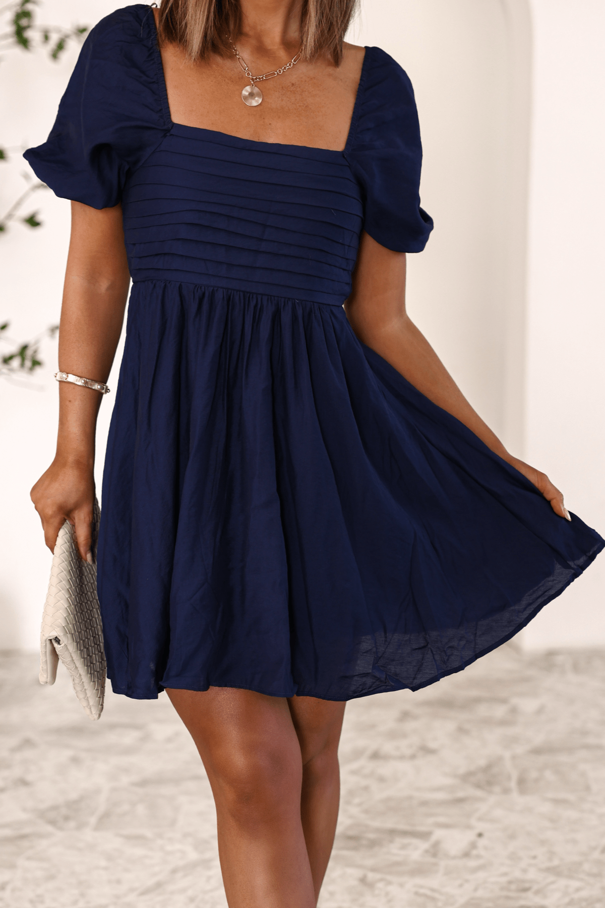 Woman in the Twilight Pleated Puff Sleeve Mini Dress-FINAL SALE holds a light clutch while standing indoors.