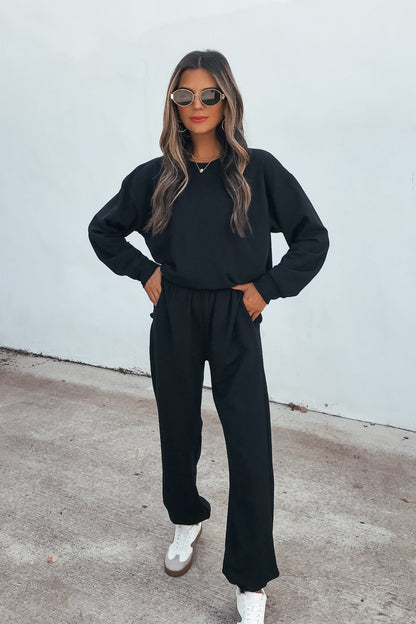 Woman in sunglasses wears a black sweater and Ultra Soft Brushed Fleece Sweatpants - Black with white sneakers by a light-colored wall.