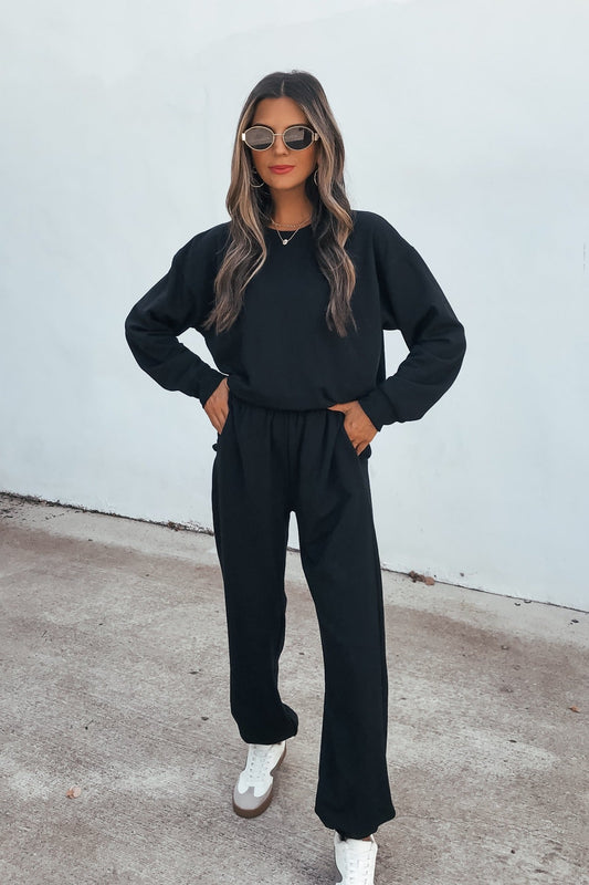 Woman in sunglasses wears a black sweater and Ultra Soft Brushed Fleece Sweatpants - Black with white sneakers by a light-colored wall.