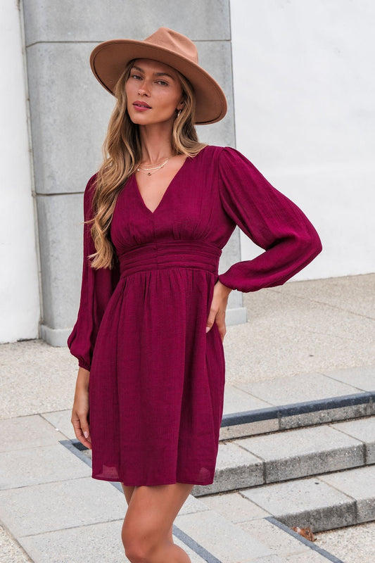 Woman in the Vanessa Burgundy Ruched Mini Dress-FINAL SALE and a tan hat poses outdoors—ideal for a fall wedding guest look.