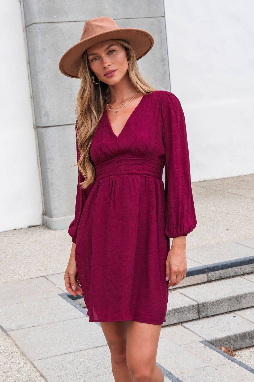 Woman in the Vanessa Burgundy Ruched Mini Dress stands outdoors on pavement, posing with one hand relaxed by her side.