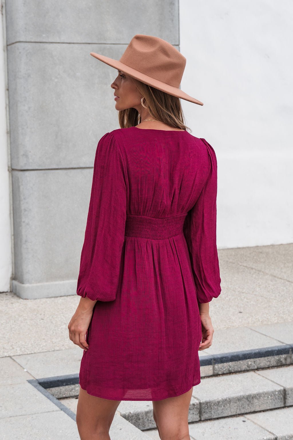 Woman in the Vanessa Burgundy Ruched Mini Dress stands outdoors in a wide-brim tan hat, facing away from the camera.
