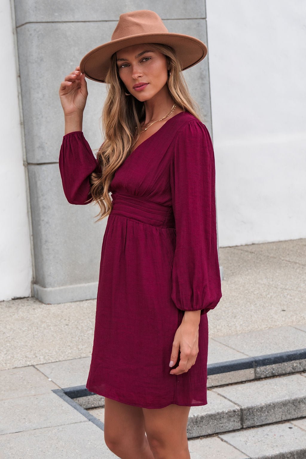 Woman wears the Vanessa Burgundy Ruched Mini Dress-FINAL SALE and tan hat, standing outdoors on a paved area, facing the camera.