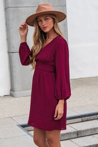 Woman wears the Vanessa Burgundy Ruched Mini Dress-FINAL SALE and tan hat, standing outdoors on a paved area, facing the camera.