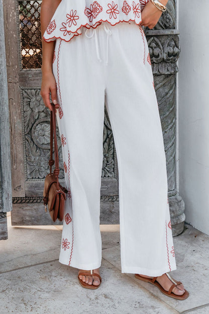 Woman in Veranda White Embroidered Pants (FINAL SALE), matching top, brown sandals, and purse by a carved wooden door.