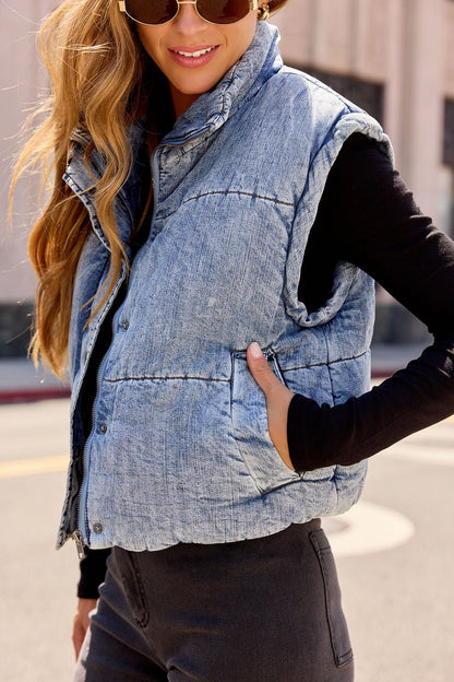 Woman smiles outdoors in a Vintage Havana Puffer Vest - Denim, layered over a black high-neck top and accessorized with sunglasses.