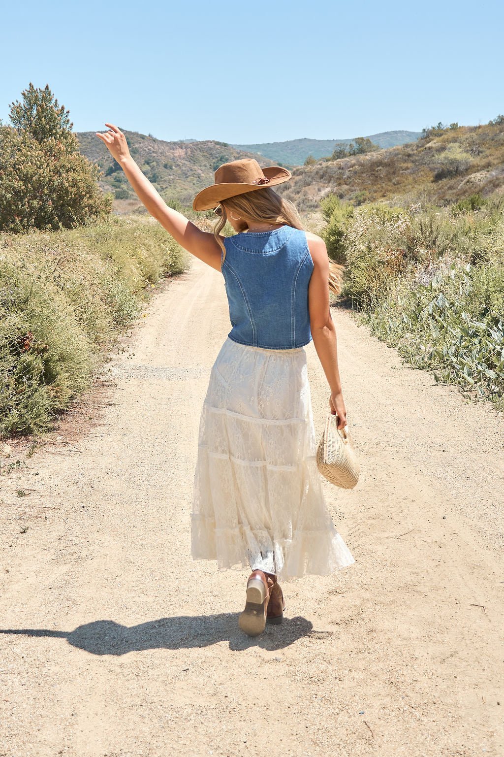 Woman in the Weston Button Front Denim Vest strolls a sunlit dirt path, pairing it with a white skirt, hat, and woven bag.
