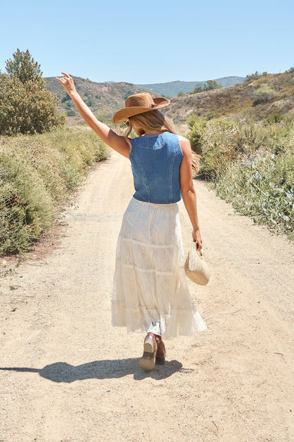 Woman in the Weston Button Front Denim Vest strolls a sunlit dirt path, pairing it with a white skirt, hat, and woven bag.