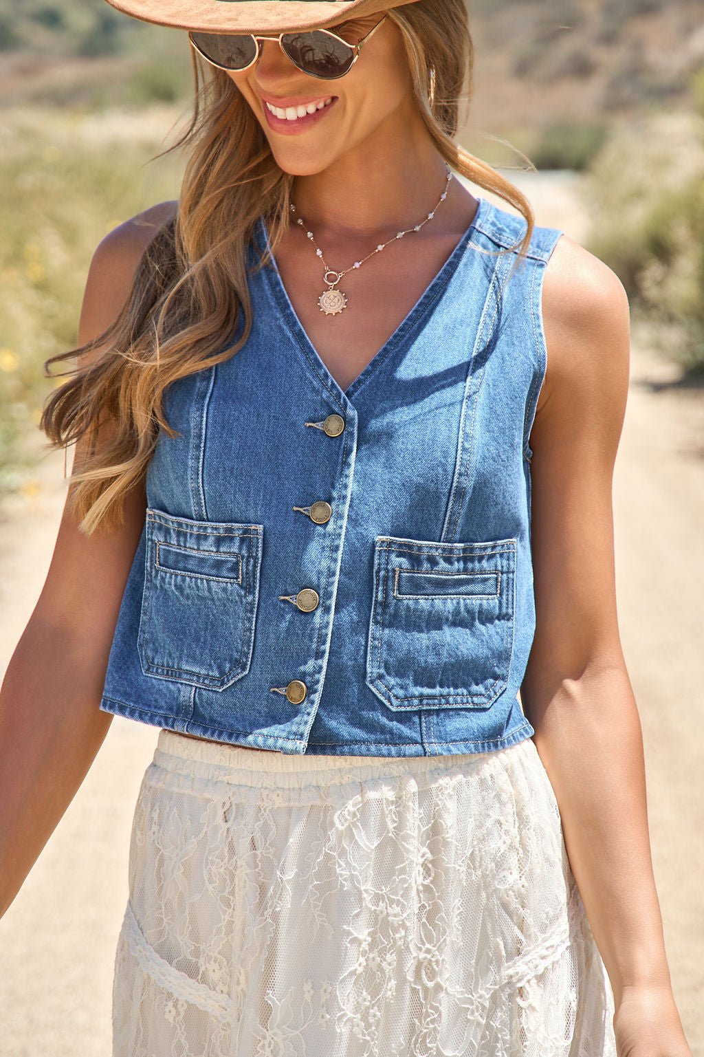 Woman wears the Weston Button Front Denim Vest, white lace skirt, sunglasses, and sunhat while smiling outdoors on a sunny path.