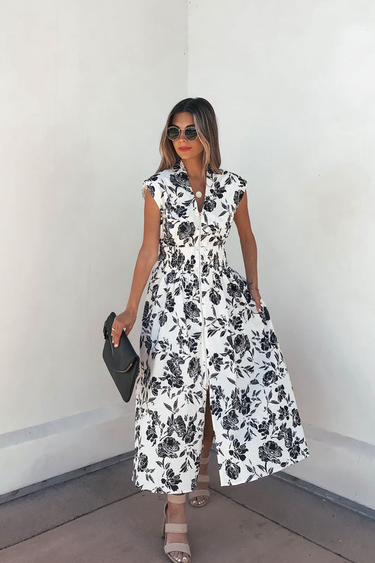 Woman in a White and Black Floral Print Smocked Midi Dress-FINAL SALE, sunglasses, black clutch, sandals, by a white wall.