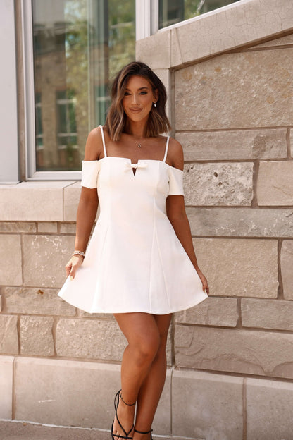 Woman in a White Cold Shoulder Flare Mini Dress - FINAL SALE stands outdoors by a stone wall, smiling and holding her dress.