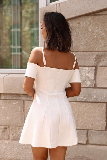 A woman in the White Cold Shoulder Flare Mini Dress - FINAL SALE stands before a stone wall, graceful and poised.