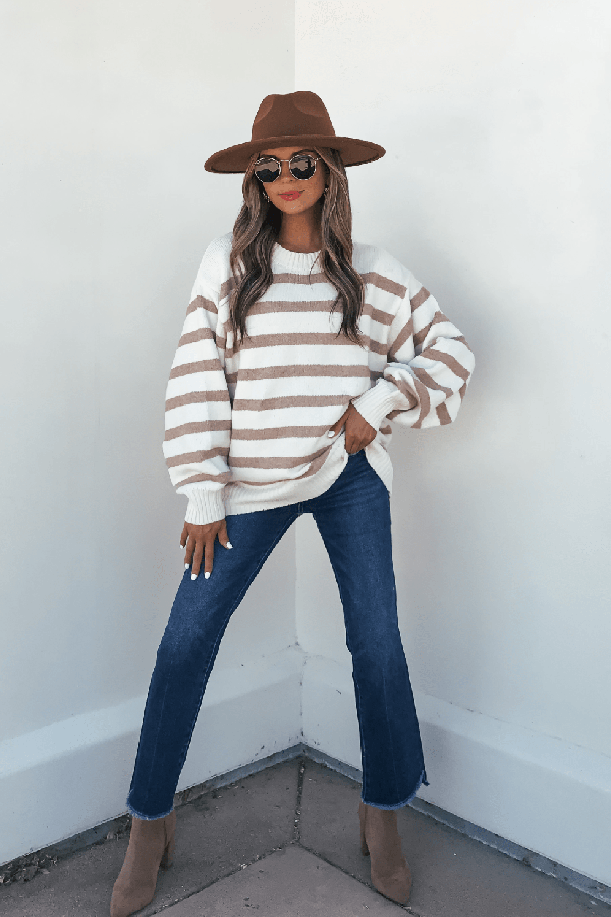 Woman in the Willow Cream Stripe Ribbed Sweater, jeans, sunglasses, boots, and brown hat poses by a white wall.
