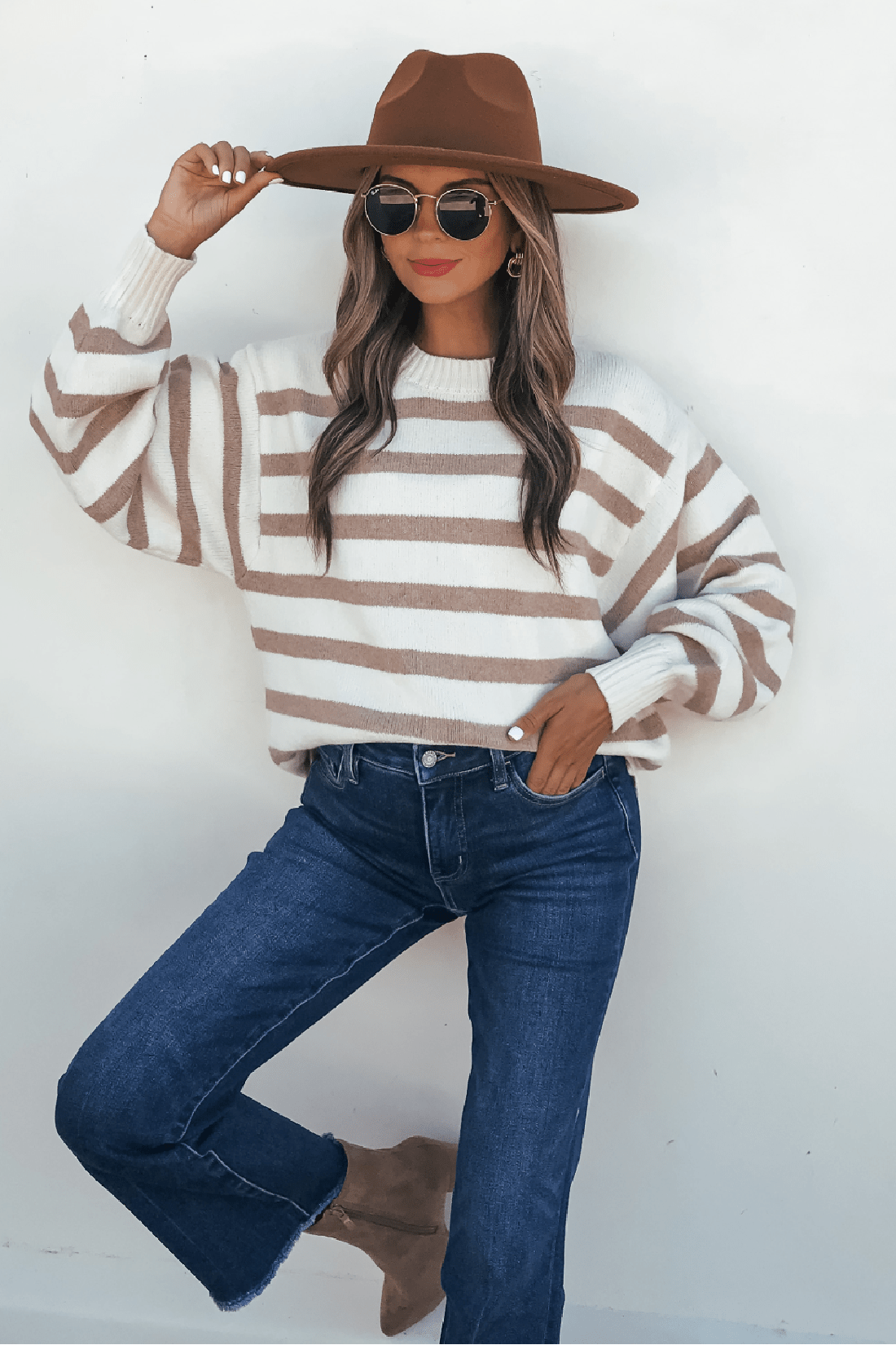 Woman in Willow Cream Stripe Ribbed Sweater, jeans, hat, and sunglasses poses by white wall with one leg bent.