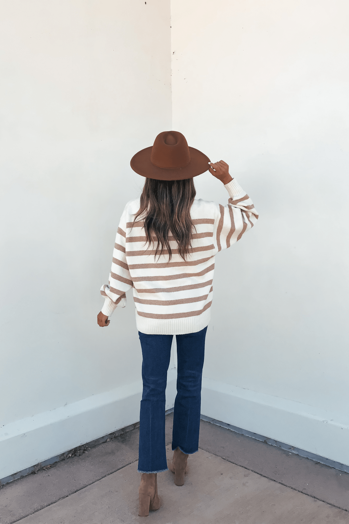 A woman in an oversized Willow Cream Stripe Ribbed Sweater, blue jeans, and boots faces a white wall with her back to the camera.