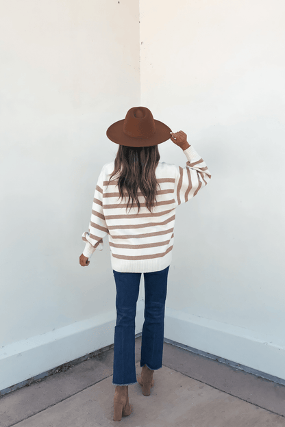 A woman in an oversized Willow Cream Stripe Ribbed Sweater, blue jeans, and boots faces a white wall with her back to the camera.