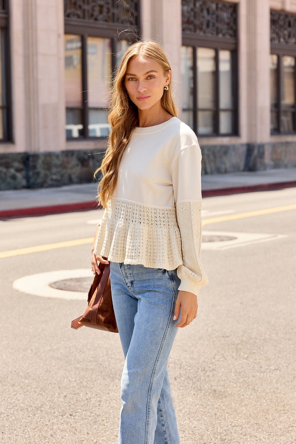 Woman with long blonde hair in the Wren Textured Sweater - Cream and jeans stands on a city street holding a brown bag, looking at the camera.