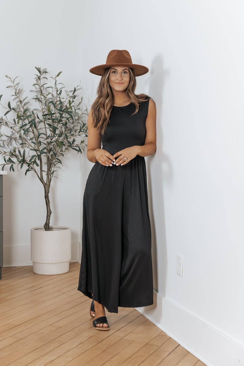 Woman in the Black Open Back Wide Leg Jumpsuit - FINAL SALE and a brown hat stands by a potted plant on a wooden floor, facing forward.