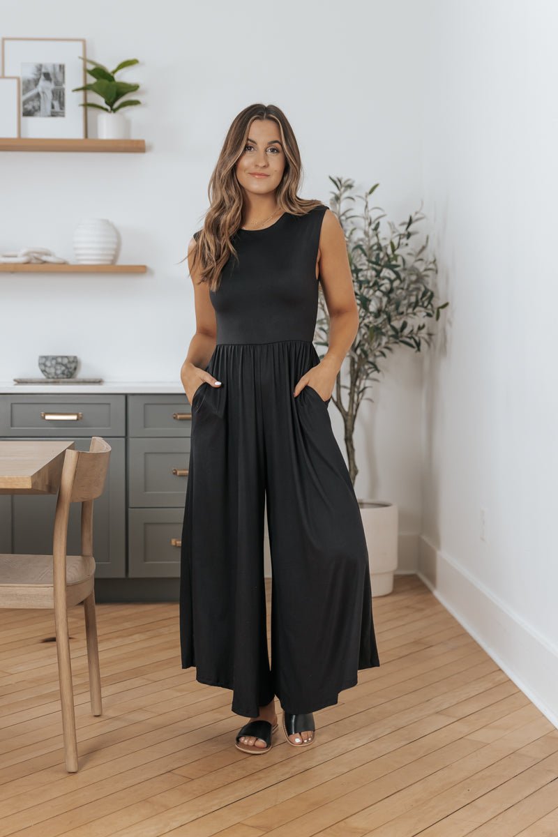 Woman models the Black Open Back Wide Leg Jumpsuit - FINAL SALE indoors beside a plant and modern furniture.