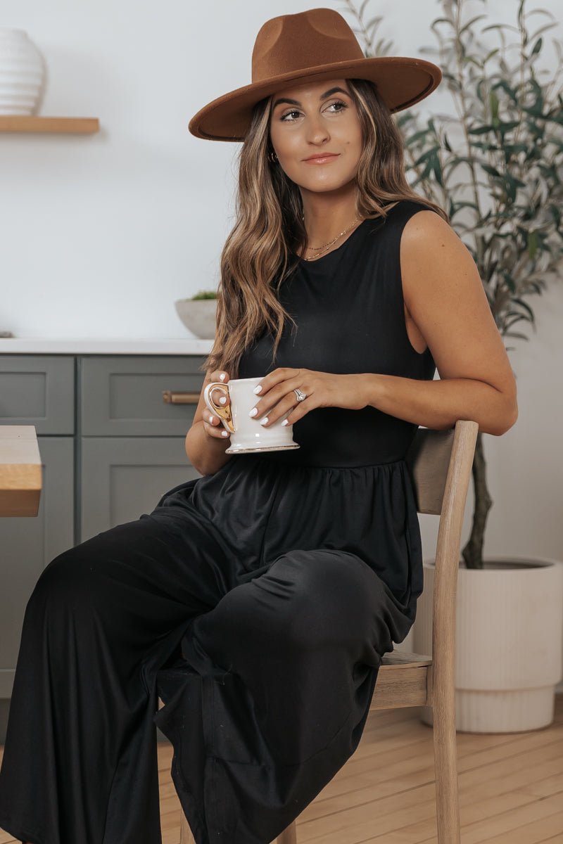 Woman wears Black Open Back Wide Leg Jumpsuit - FINAL SALE, sitting with a mug and brown hat in a modern kitchen with plants.