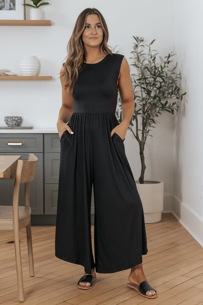 Woman indoors in a Black Open Back Wide Leg Jumpsuit - FINAL SALE, black sandals, hands in pockets on a light wood floor.