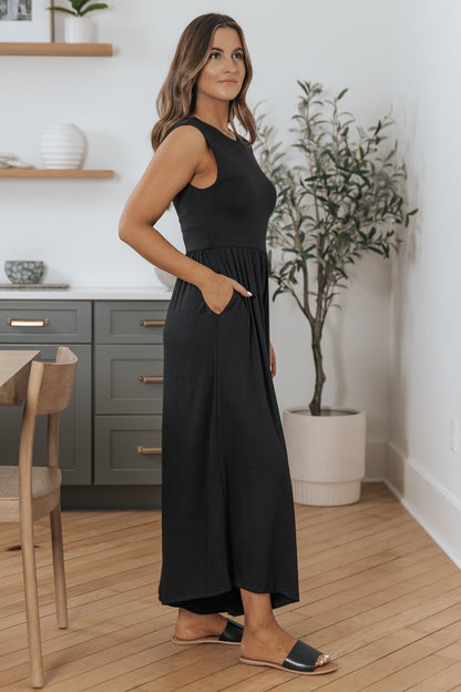 Woman wears Black Open Back Wide Leg Jumpsuit - FINAL SALE, standing indoors with plants and modern decor in the background.