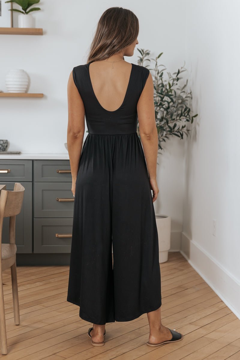 Woman models the Black Open Back Wide Leg Jumpsuit - FINAL SALE, indoors on wooden floors, facing away from the camera.